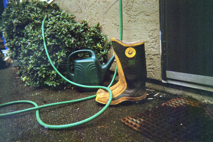A pair of black and yellow rain boots resting next to a dark green watering can. The boots and water can and resting between a doormat and a shrub.