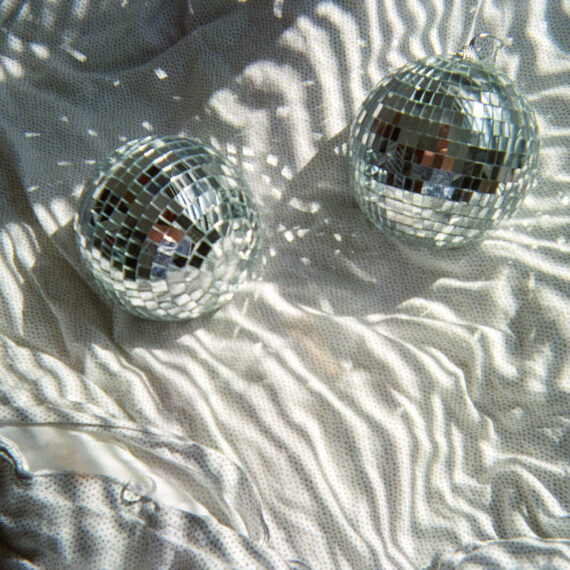 Two mini silver disco balls on a white bedsheet. There is sunlight shining on the disco balls that are causing refractions of light to show on the bedsheet.
