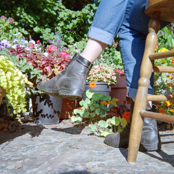 A person wearing jeans and black boots sitting cross legged on a wooden chair. In front of them is a variety of potted plants and flowers. The camera only shows the legs of the person and one leg of the wooden chair.
