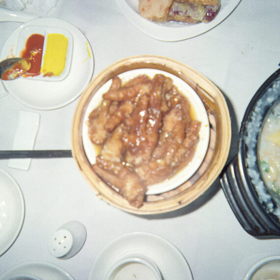 Colour photo of a spread of food on a white table cloth. There are cups of tea, plates of sauce, and pots of congee and duck feet.