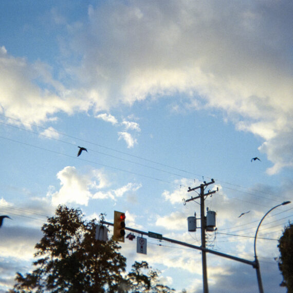 A photo of a light blue sky with a few clouds scattered around. The photo is taken at an intersection with a red light, a power line, trees, and a few birds flying by.