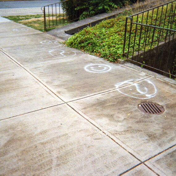 White smiley faces spray painted along a sidewalk. Next the sidewalk is a railing fence and a concrete ramp overgrown with ivy.