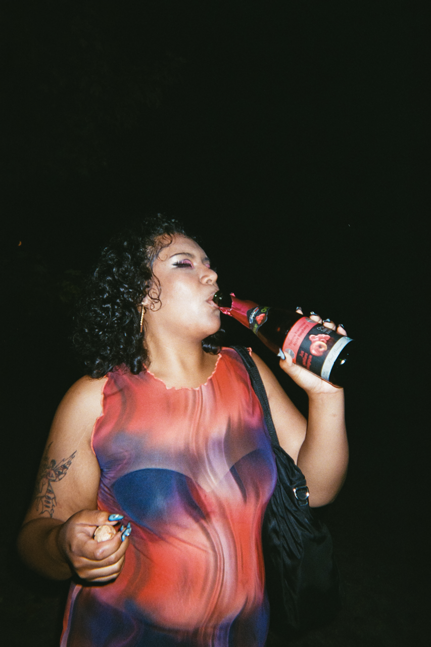 A person drinking from a large sparkling juice bottle, their closed eyes showing peachy pink eyeshadow that matches their dress and the label on the bottle. They have shoulder-length black curly hair and a black purse under one arm, blending into the dark night behind them.