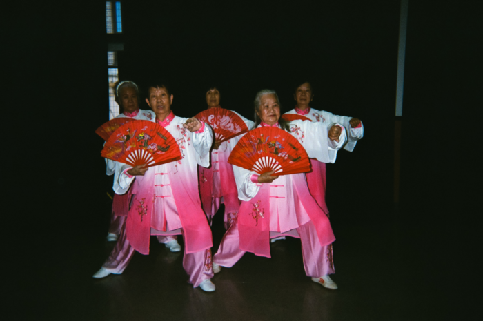 Five senior Chinese women in white and pink tai chi uniform hold their large red half-circle fans in front of their torsos as they hold an arm to the side mid tai chi form.