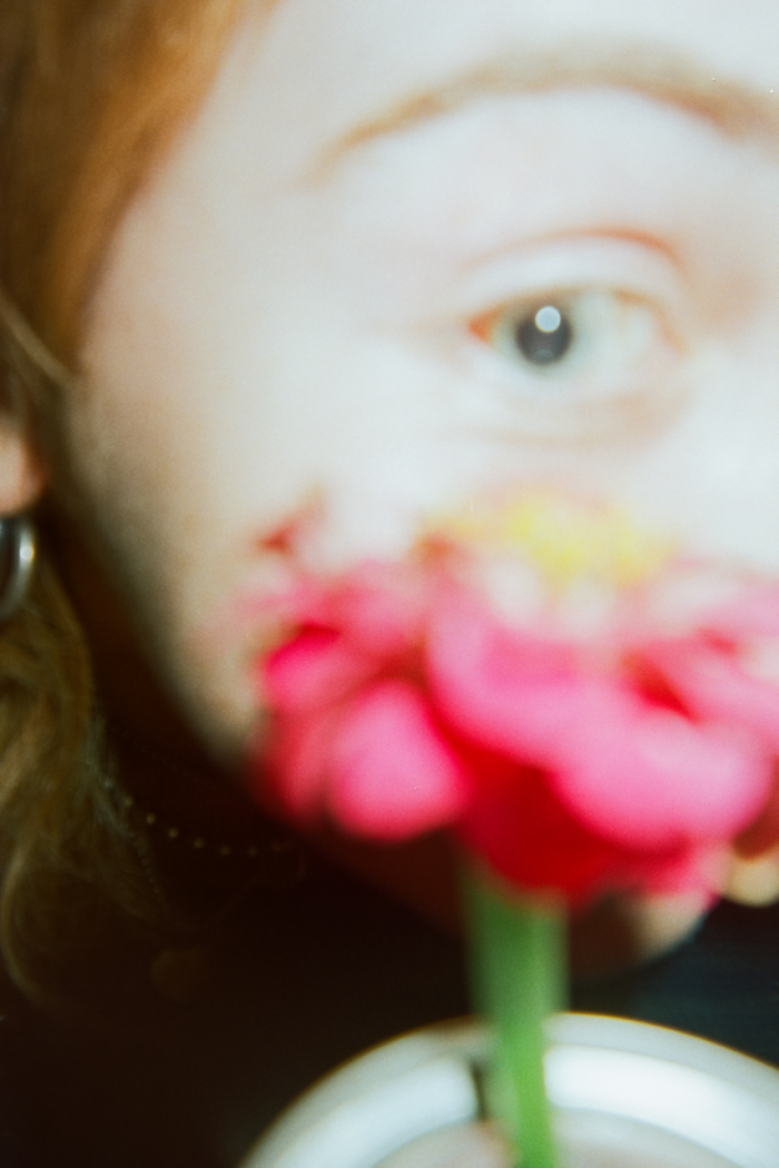Out of focus close-up of a person's eye with a many-petaled pink flower held up to the person's cheek hiding the rest of their face. In the camera's flash, their skin is pale, their pupil large, and their hair tucked back a copper color.