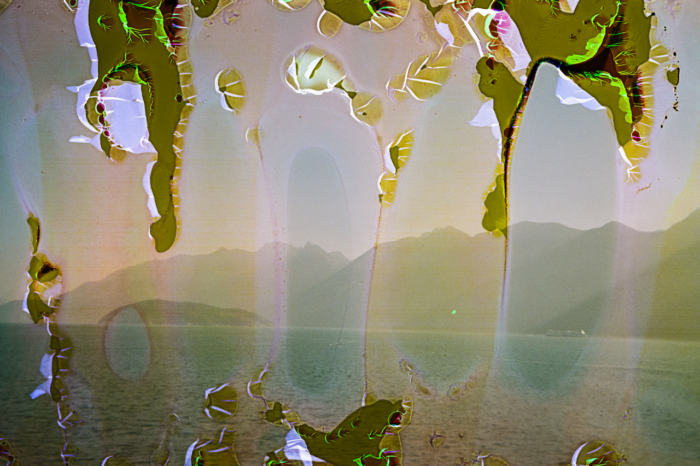 A horizon of tall mountains beneath a pale sky, wide blue water of the ocean at their base. Out on the water is a small, wooded island and a ferry crossing the water. The photo shows salt water damage to the film, with large blotches of chromatic abberations along its top and bottom.