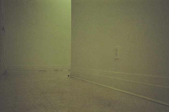 A photo taken low to the ground of a hallway. The floor, vinyl moulding, and walls are nearly the same off-white, blending them together. Along the wall that stops mid-frame with a sharp turn in the hall, a single eletrical outlet also blends into the wall.