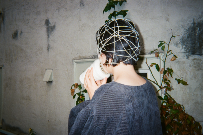 A person turned away from the camera, holding a cone of thread to their face, a web of white thread wrapped is around their head over short black hair. They stand outside, by a bush of half dead leaves and an off-white plaster wall. They wear a dark, long sleeve, corduroy shirt.