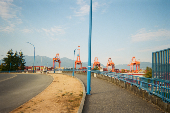Five bright orange shipping container cranes against a mountain horizon and blue sky seen from the sidewalk of a raised road that curves away to the left. The rail and streetlamps of the road are a bright blue, the post of the nearest streetlamp is centre-frame.