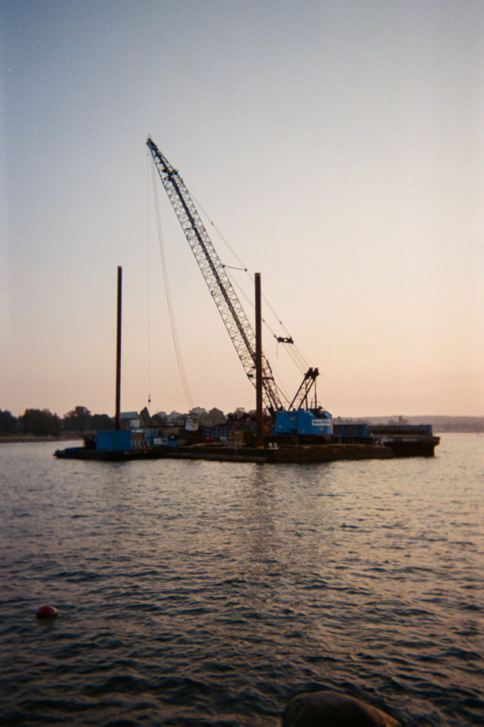 A barge floating on dark, choppy water reflecting the pale sky of a setting sun. The low, flat barge has two tall poles reaching into the sky alongside a much taller crane arm that long cables hang from.
