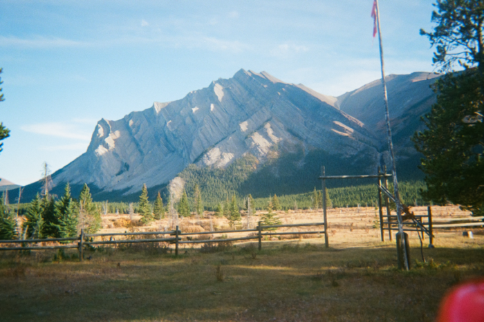 Past a wood log fence and flat dry grass with short pine trees, a tall mountain of pale grey rock sits mostly shaded from the sun. The mountain is composed of sweeping lines of strata near vertical. In front of the wooden fence is a flag pole flying the Canadian flag.