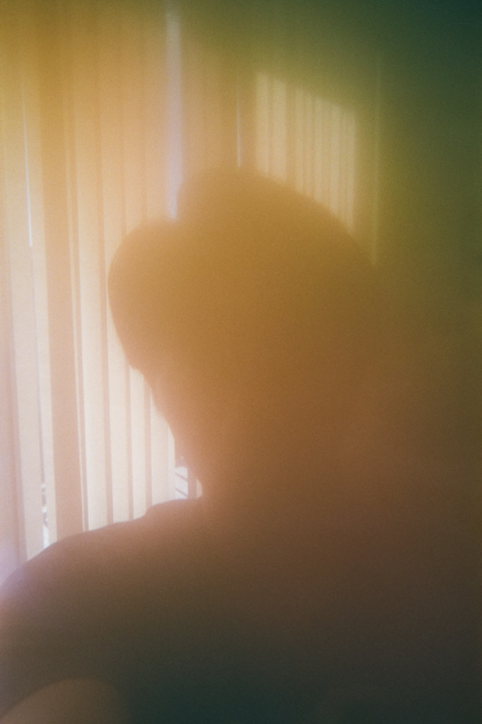 The silhouette of a person's head and shoulders in a soft warm hazy light pouring through a large window with vertical blinds.