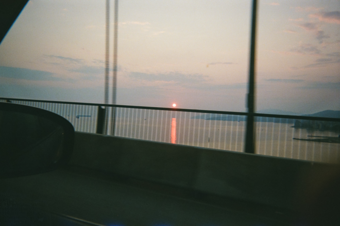 The view of ocean and a low sun from the passenger's seat of a car as it crosses a bridge. Just above the rail of the bridge, the sun is a small orange orb, casting a line of orange on the water below. The sky and distant mountains of shoreline are in a soft, cloudy haze.